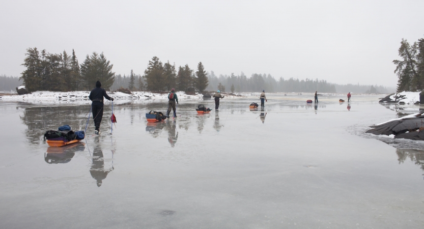 a line of cross country skiers, each pulling small sleds, make their way across a frozen lake